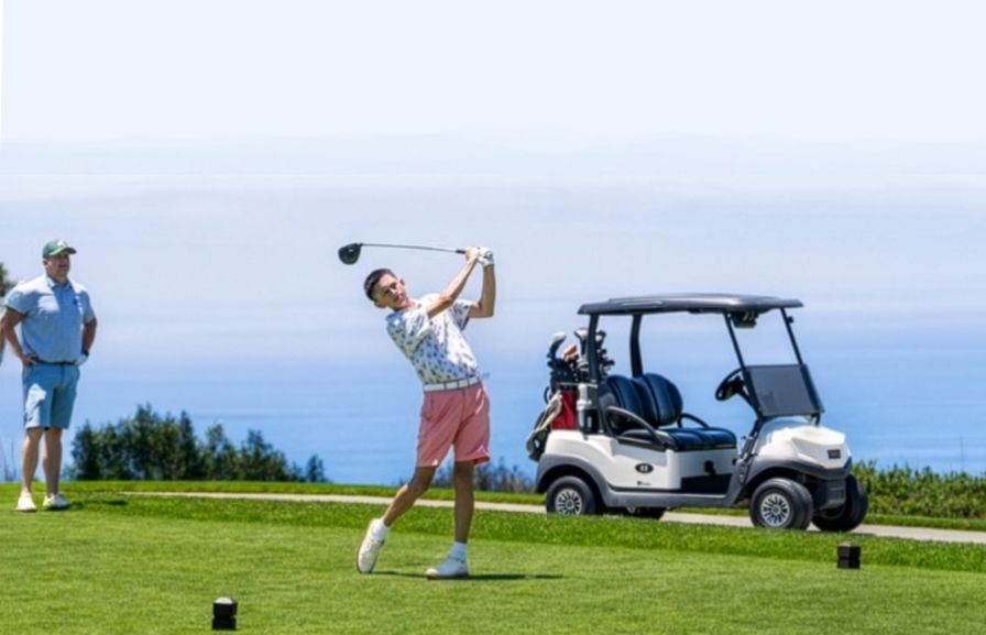 Orion driving off an ocean-view tee while his dad watches beside the cart.